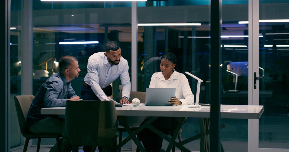 Three colleagues in a glass-walled boardroom collaborate around a table at night, with a woman using a laptop while two men discuss nearby.
