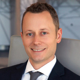 Professional headshot of a smiling man in a dark suit and tie, seated in an office setting.