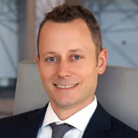 Professional headshot of a smiling man in a dark suit and tie, seated in an office setting.